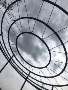 Cover image for “In your car, you track the distance of lost homes” by Purvi Shah, featuring a geometric metal structure silhouetted against a cloudy gray sky.