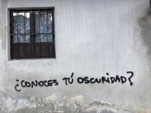 Cover image for "On Healing" by Courtney Ng, featuring a barred window and a grey concrete wall with the words ‘¿Conoces tu oscuridad?’ spray-painted on it.