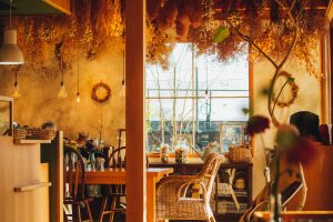 Cover image for “To the Obāsan at the Breakfast Café” by Lauren Mariko Scherr, featuring tables and chairs in a cafe decorated in warm fall colors.